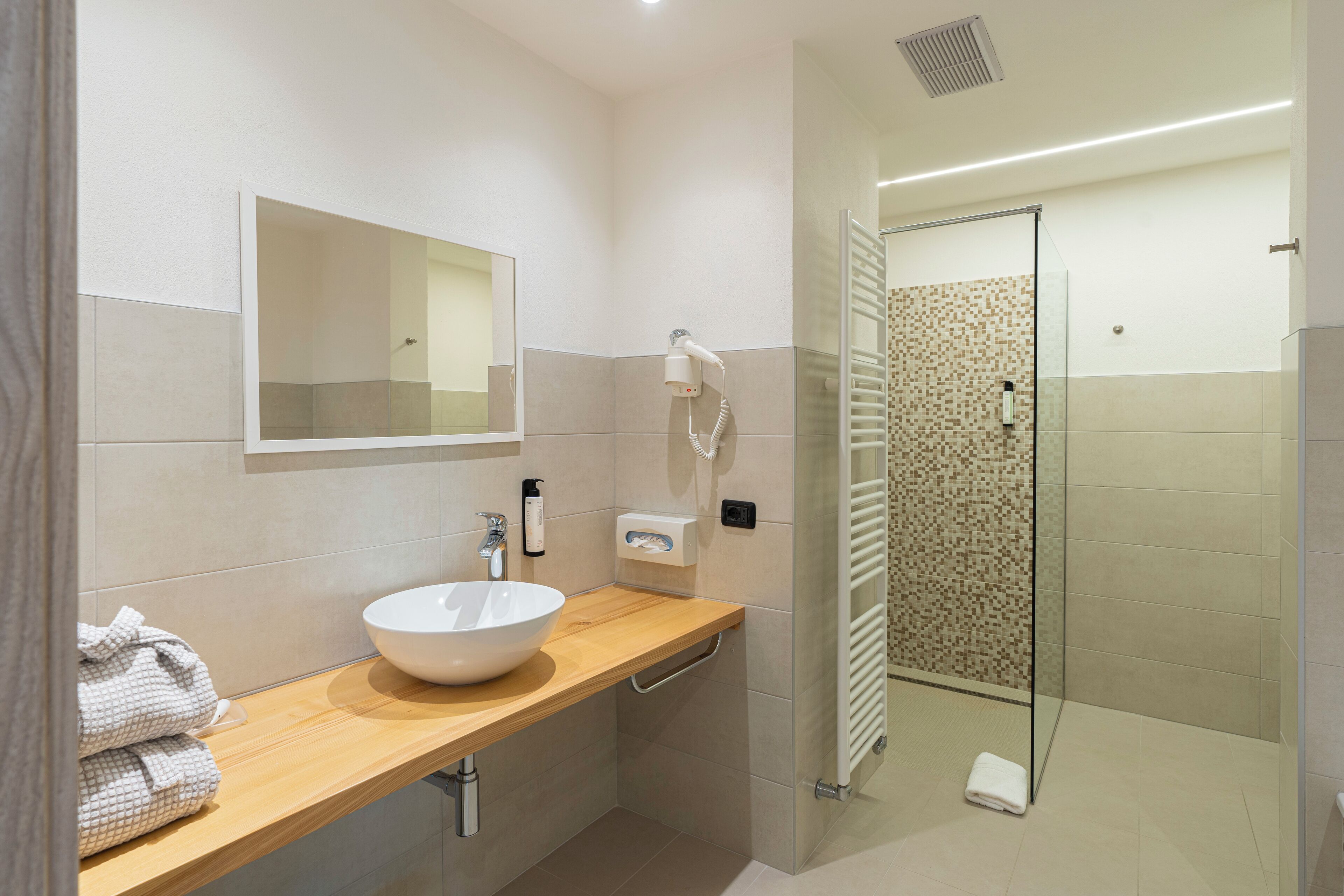 A modern bathroom with a wooden vanity, a large mirror, a white sink, a handheld hair dryer, and a glass shower enclosure.
