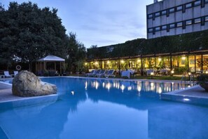 Una piscina con sillas de descanso, una formación rocosa y un edificio con ventanas iluminadas al fondo.
