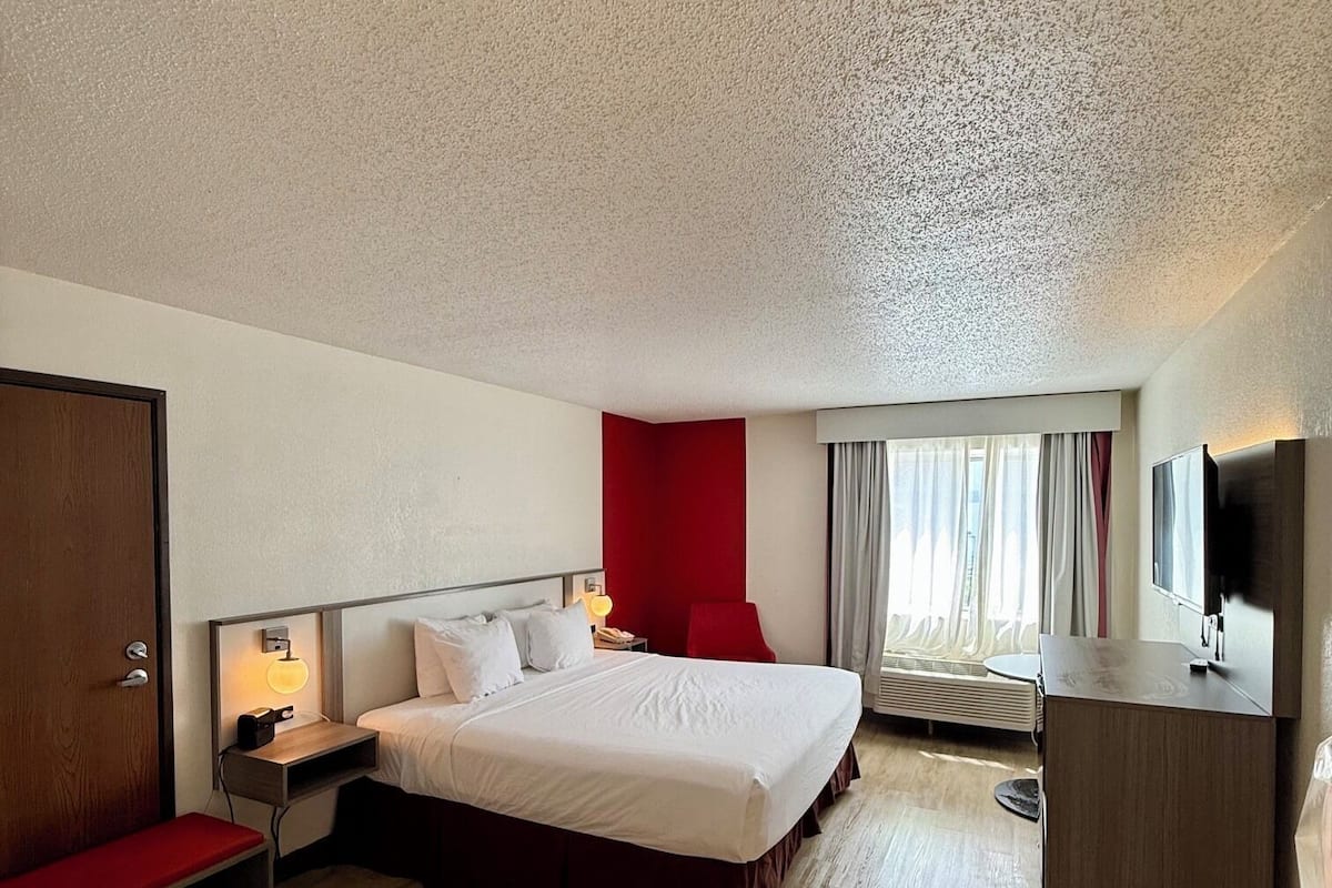A hotel room with a large bed, bedside tables, a desk, a TV, and a red accent wall.