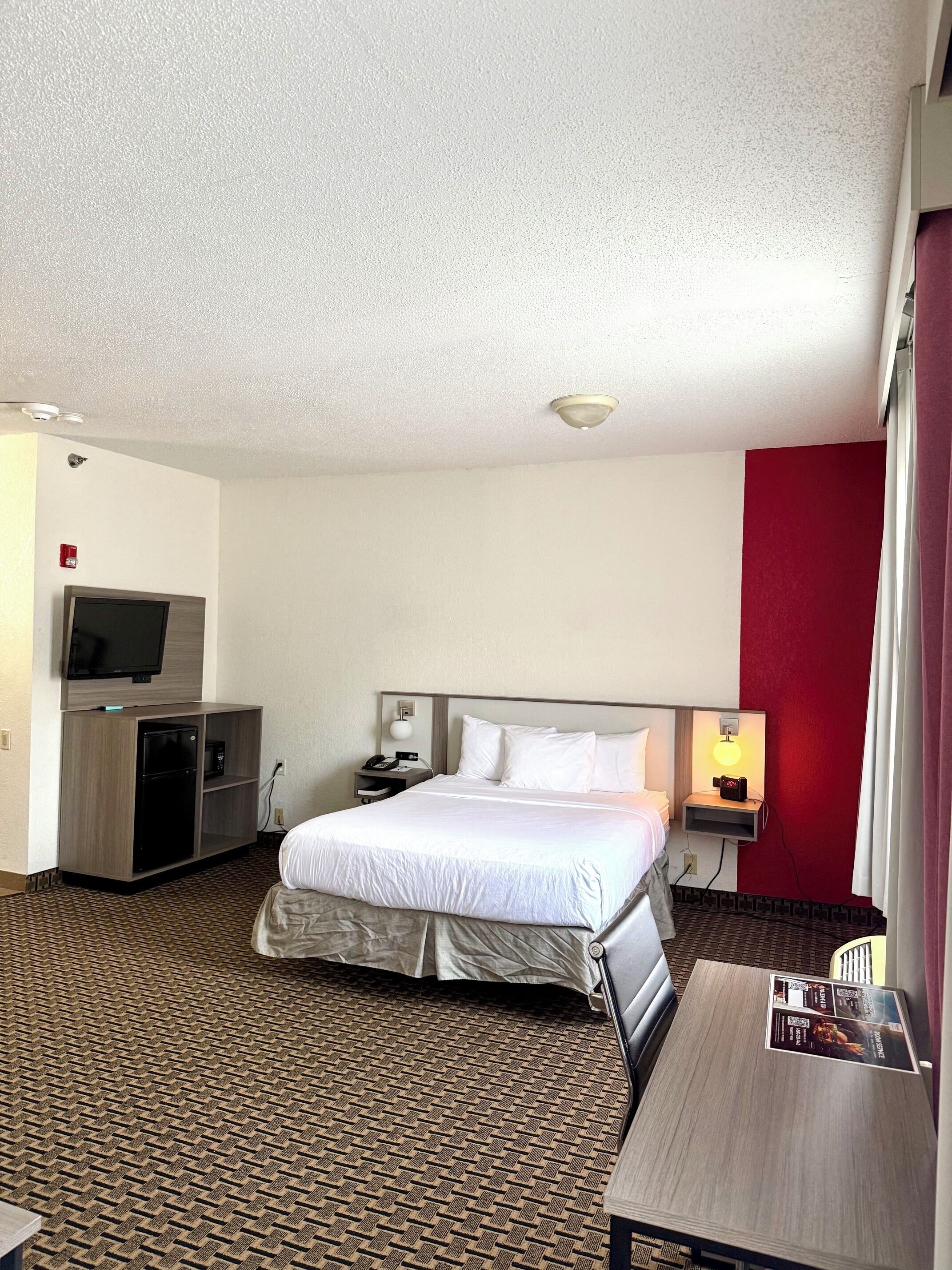 A hotel room with a bed, a TV, a minibar, a desk, and a red accent wall.