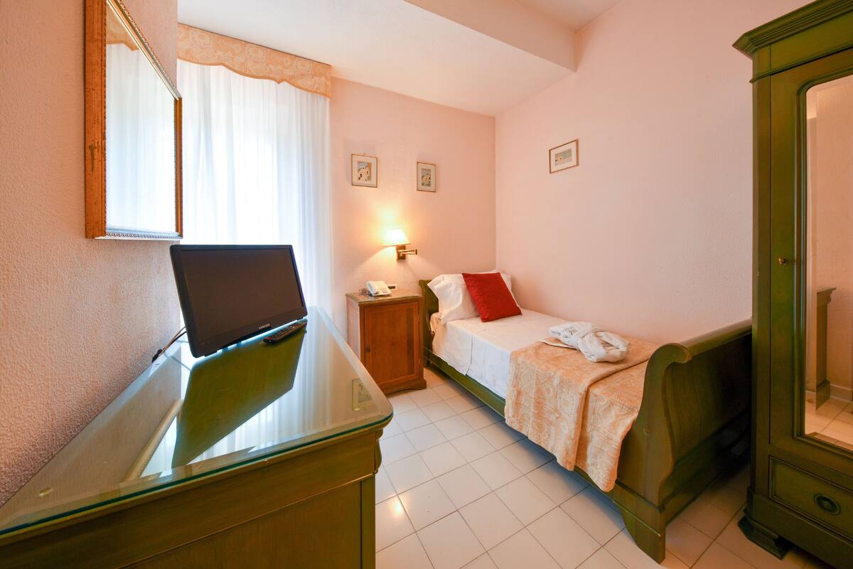 A hotel room with a bed, a bedside table, a desk with a computer, a lamp, and a mirror.
