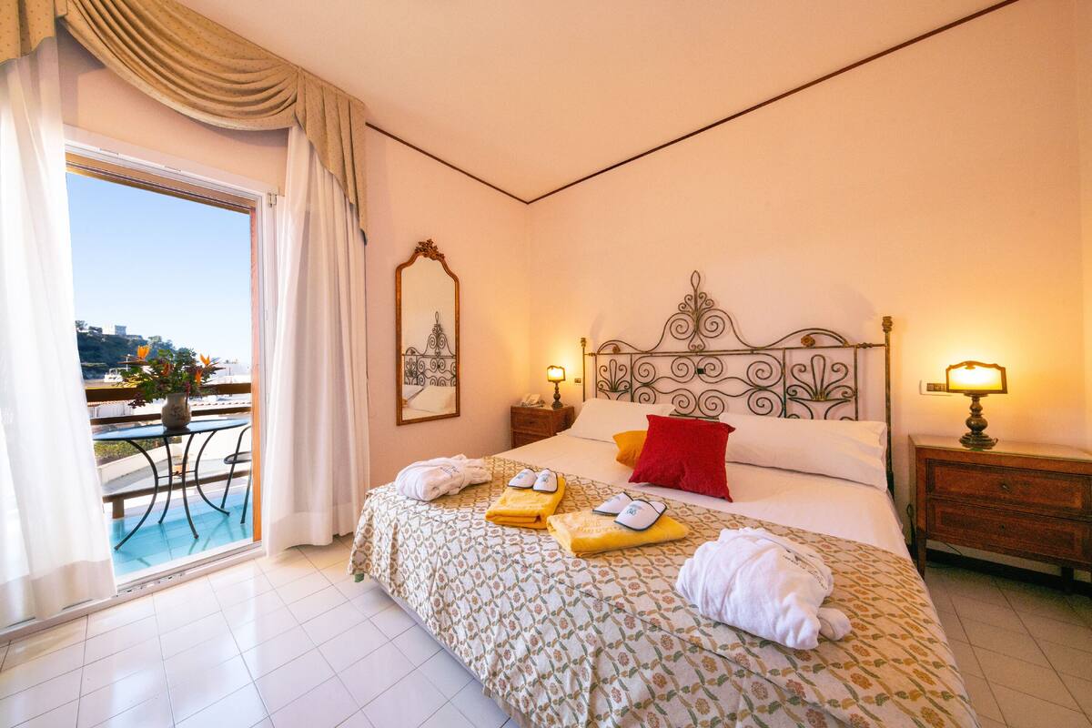 A hotel room with a bed, two bedside tables, a window with curtains, a balcony, and a view of the city.