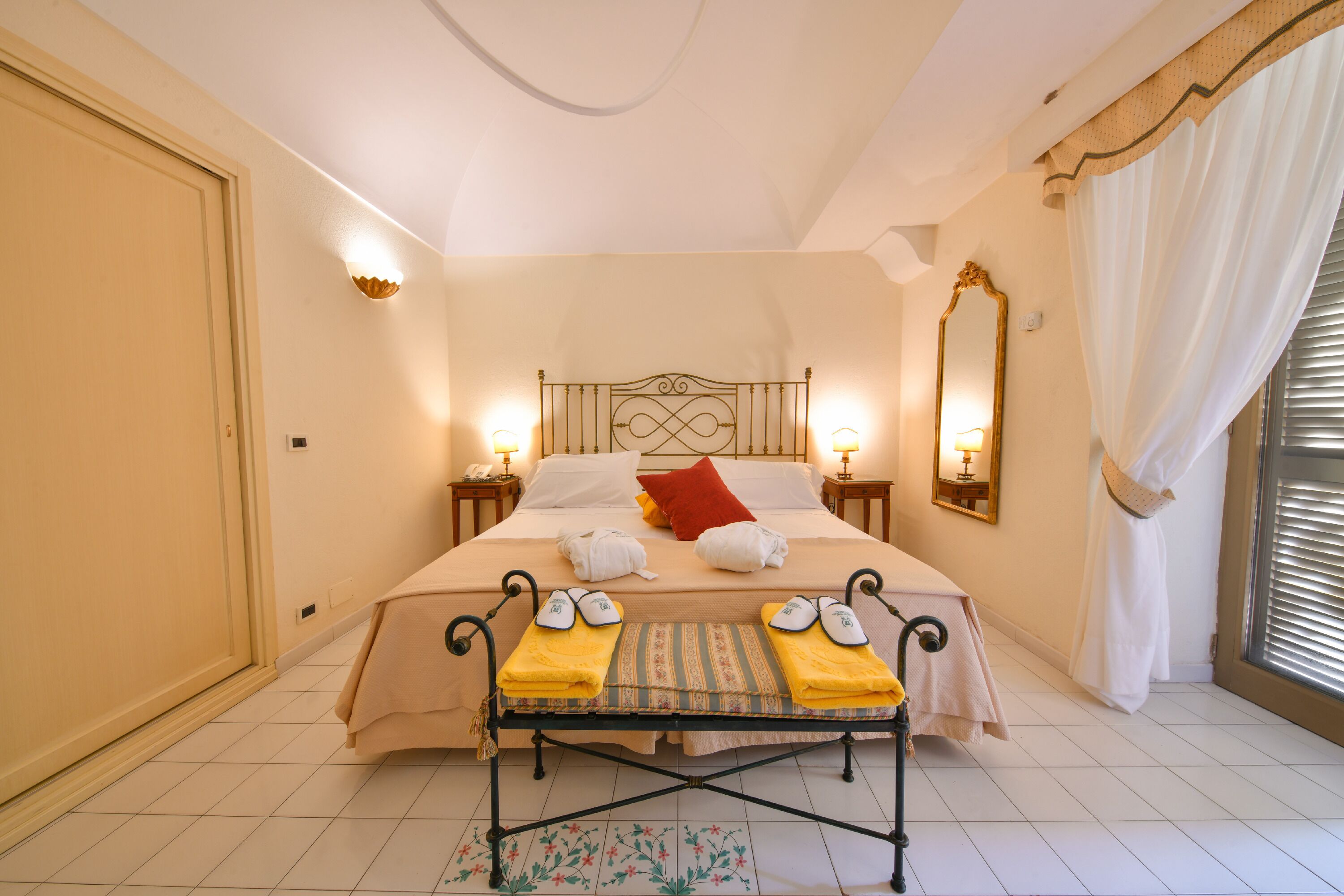 A hotel room with a bed, bedside lamps, a mirror, a window with curtains, and a small table with towels and shoes.