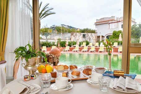 A table with breakfast items, a vase with flowers, and a view of a pool and chairs.