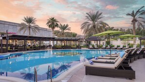 A swimming pool with lounge chairs and umbrellas, surrounded by palm trees and a building with outdoor seating.