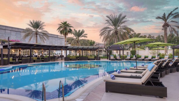 A swimming pool with lounge chairs and umbrellas, surrounded by palm trees and a building with outdoor seating. - Radisson Blu Hotel, Muscat (Muscat)
