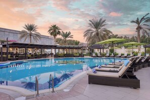 A swimming pool with lounge chairs and umbrellas, surrounded by palm trees and a building with outdoor seating.