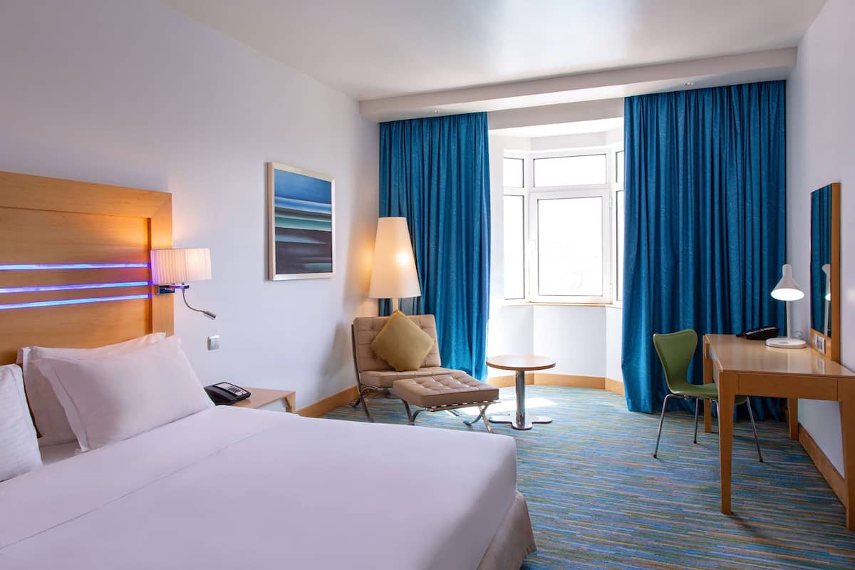 A hotel room with a large bed, a desk, a chair, a small table, a lamp, a phone, and a painting on the wall.