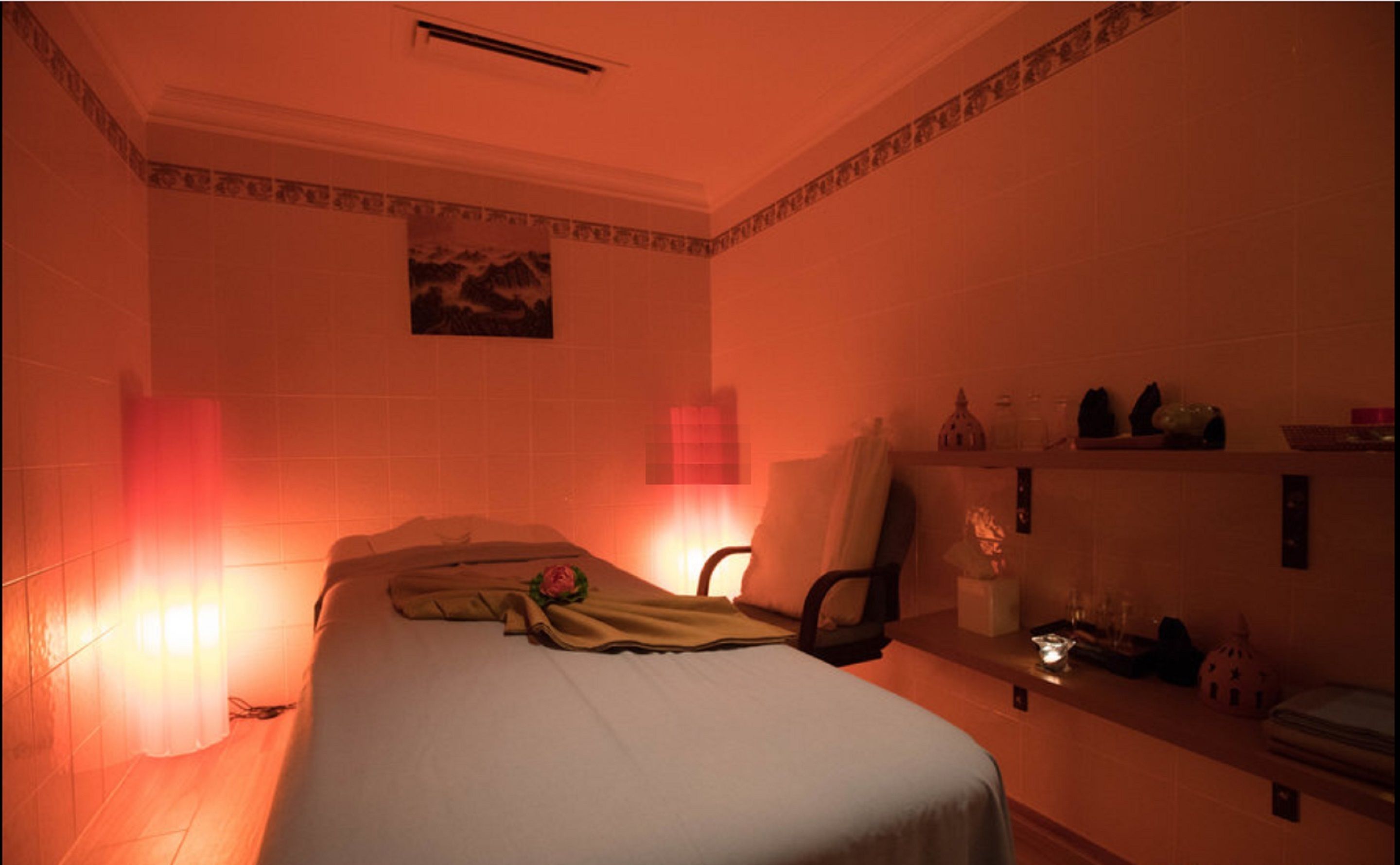Una habitación tenuemente iluminada con una cama, una silla, una mesita con objetos y un estante en la pared.
