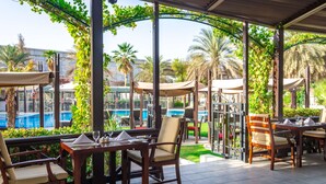 An outdoor dining area with tables and chairs, overlooking a pool and palm trees. - Radisson Blu Hotel, Muscat (Muscat)