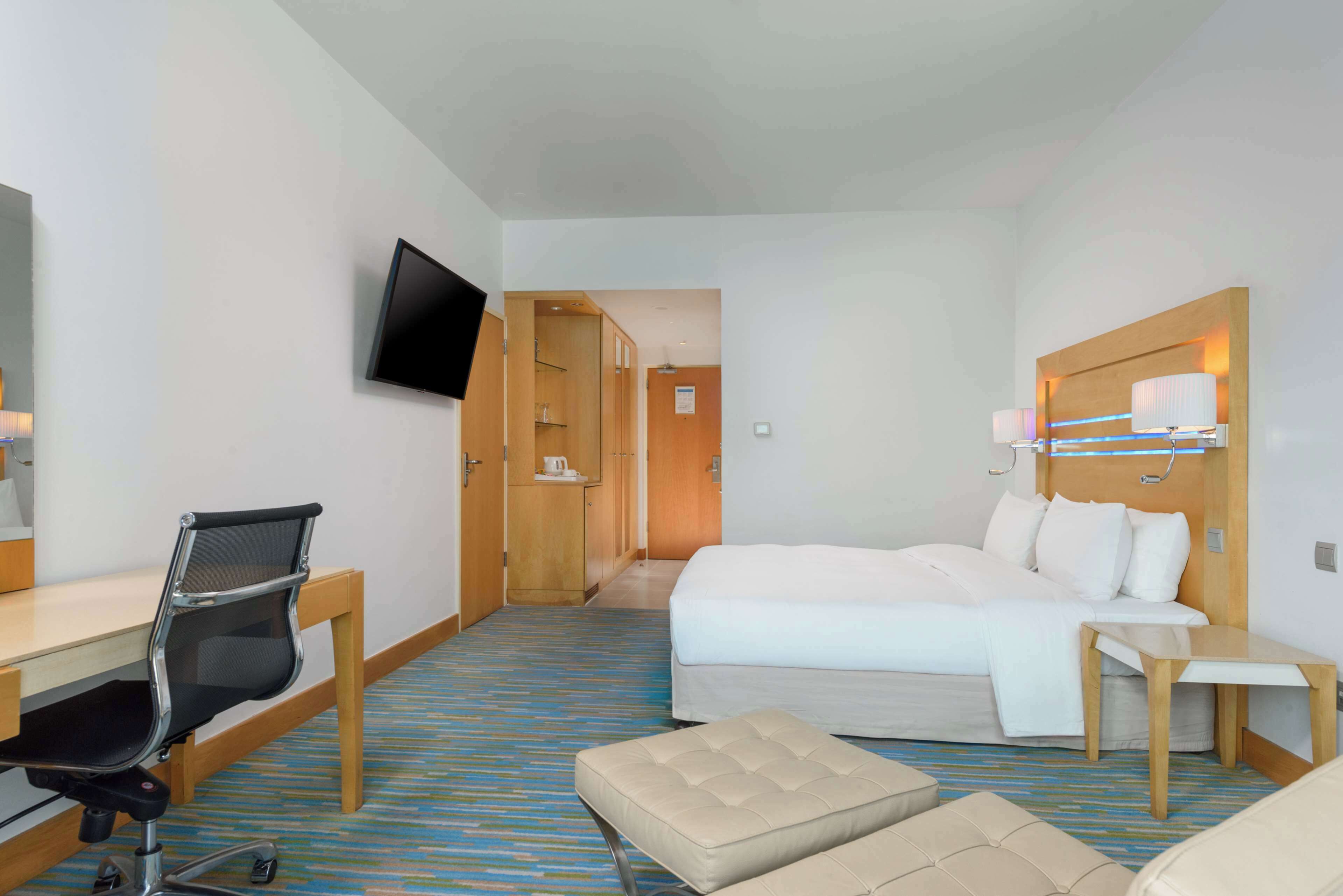 A hotel room with a bed, a desk with a chair, a television mounted on the wall, and a bedside table with a lamp.