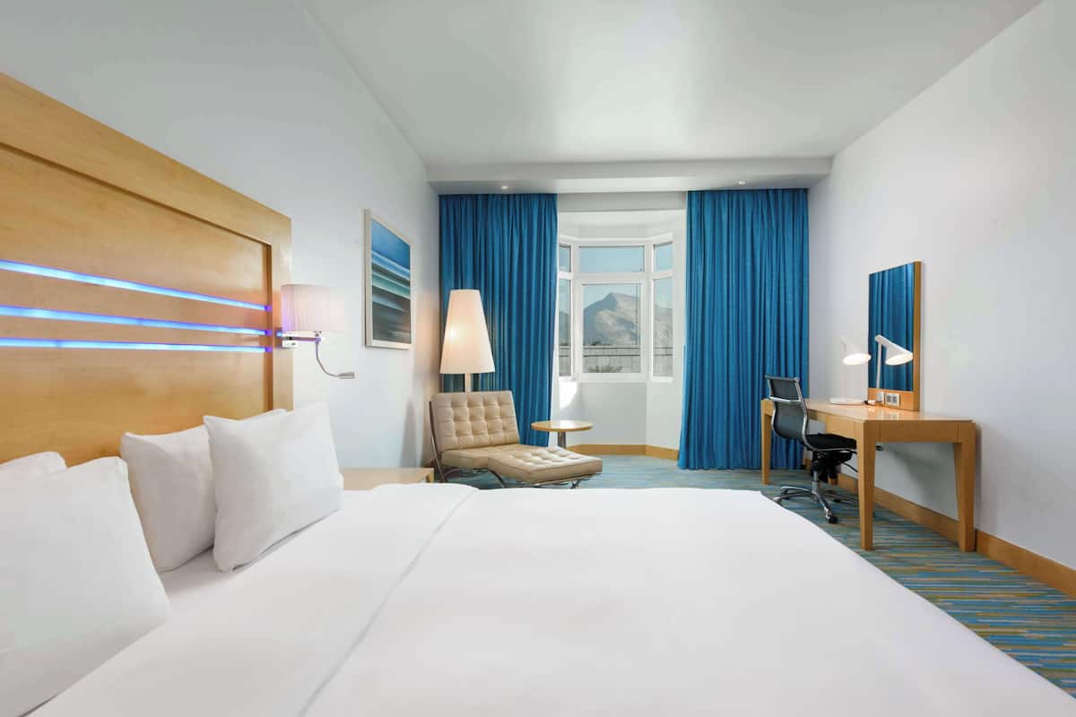 A hotel room with a large bed, a desk, a chair, and a view of a mountain through a window.