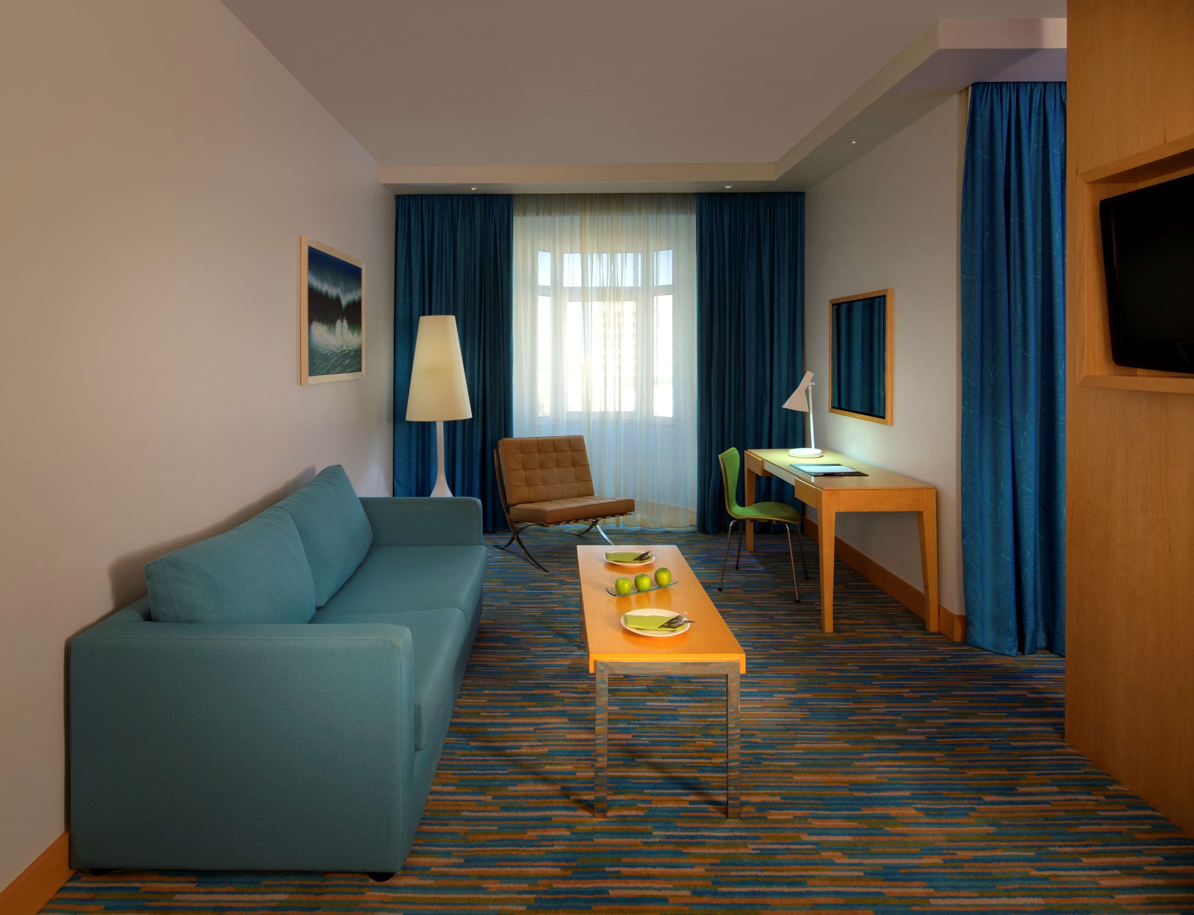 A hotel room with a blue sofa, a desk with a chair, a lamp, and a television.