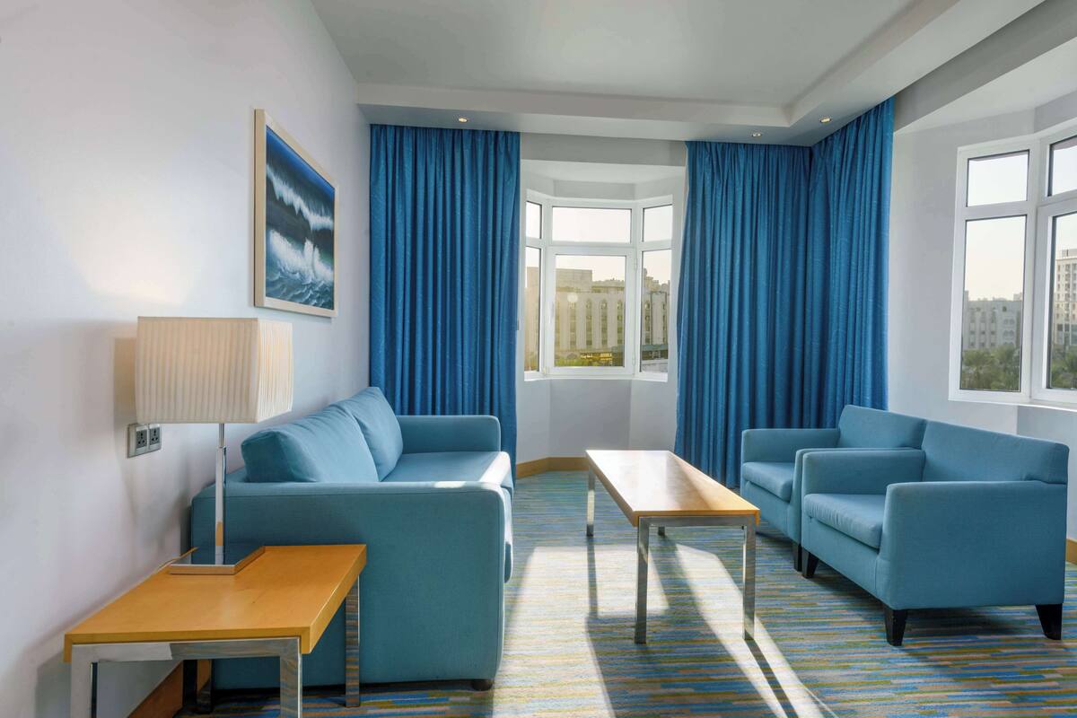 A hotel room with a blue sofa, a wooden table, and a view of the city through a bay window.