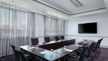 Un salón de conferencias con una mesa larga, sillas, un televisor de pantalla plana y ventanas amplias con cortinas translúcidas.