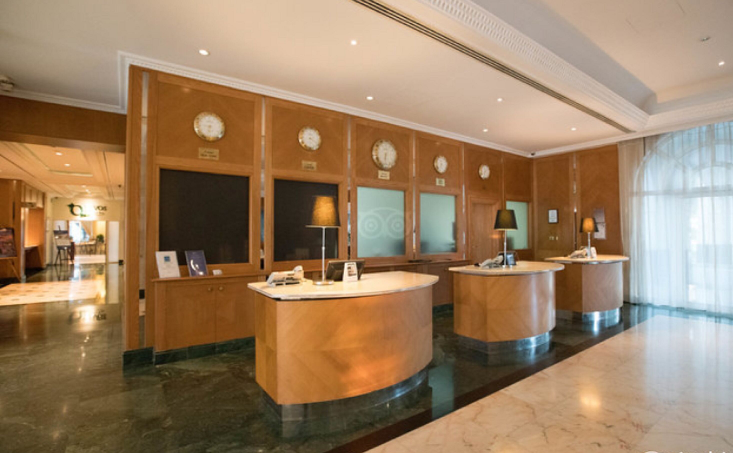 Un vestíbulo de hotel con tres mostradores de recepción, piso de mármol y amplias ventanas.