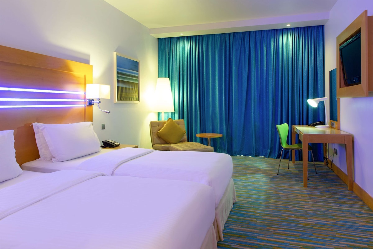 A hotel room with two beds, a desk, a chair, a TV, and a large window with curtains.