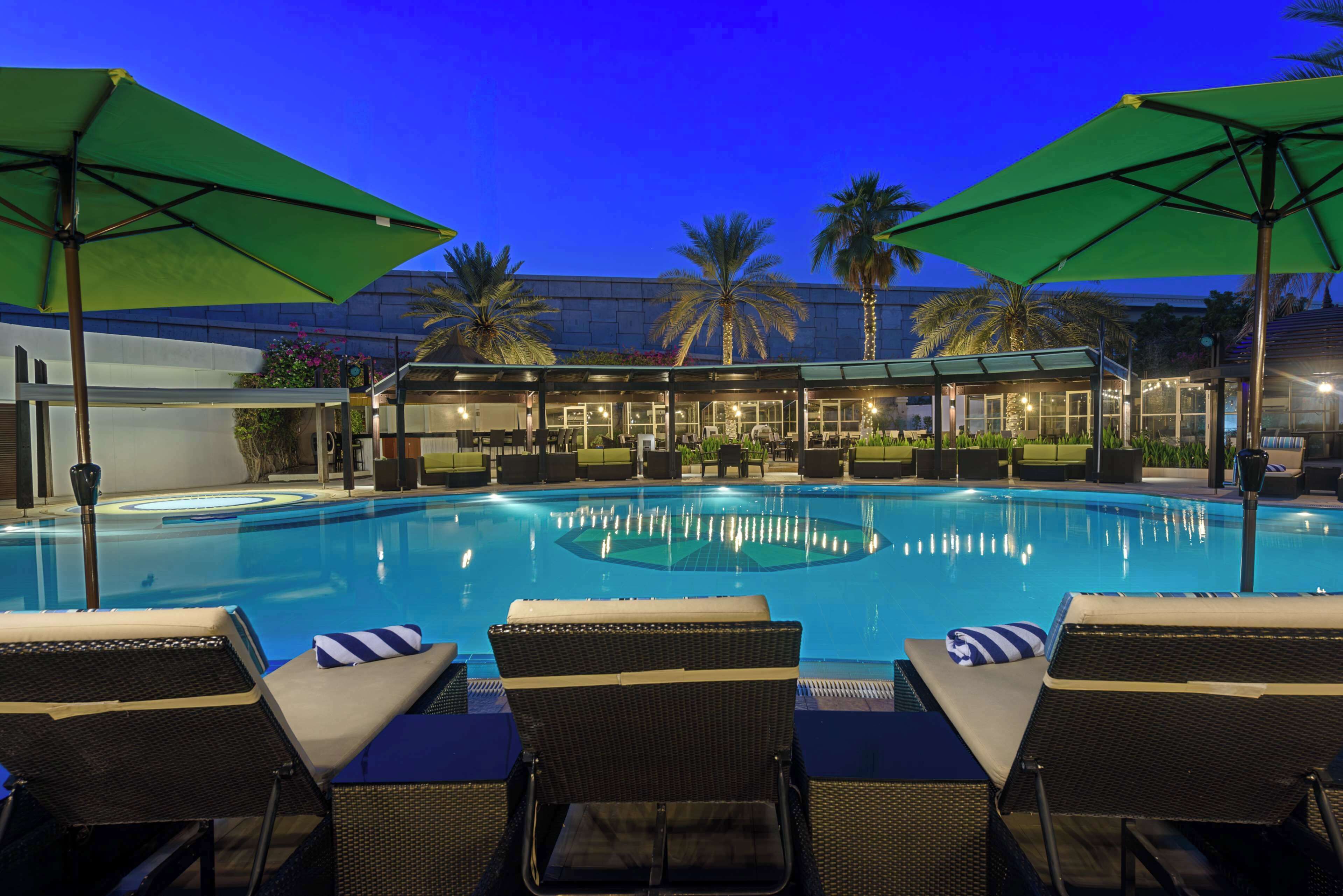 A swimming pool with lounge chairs and umbrellas, surrounded by palm trees and a building with glass doors.