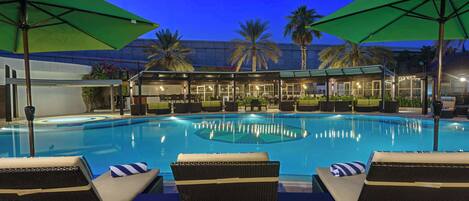 A swimming pool with lounge chairs and umbrellas, surrounded by palm trees and a building with glass doors.
