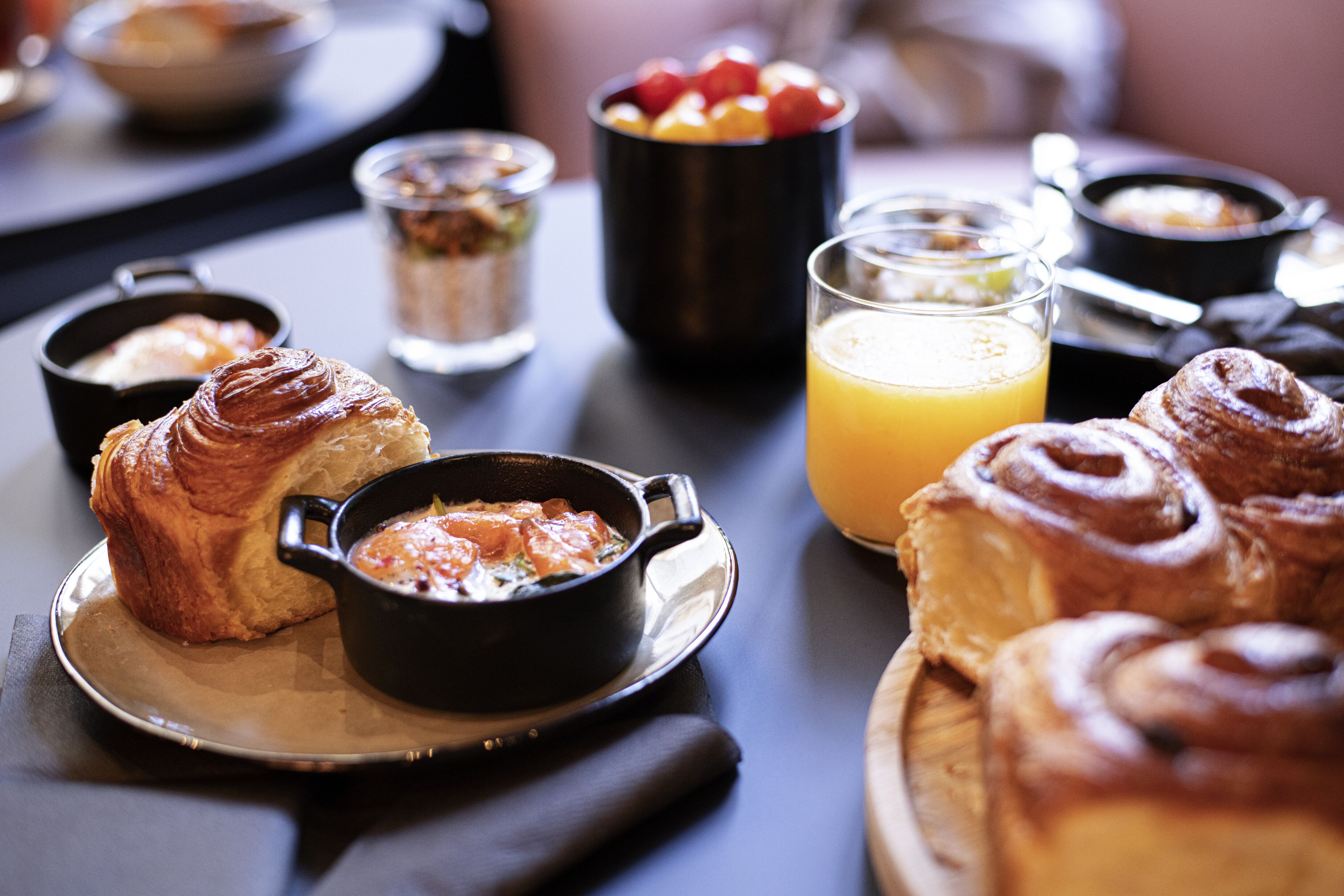 Une table dressée pour le petit-déjeuner avec des croissants, un bol de soupe, un verre de jus d’orange et un saladier de fruits.