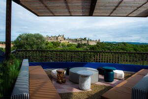 Terraço no telhado com vista para um castelo histórico, mobiliado com sofás listrados, pufes redondos e uma mesa pequena.