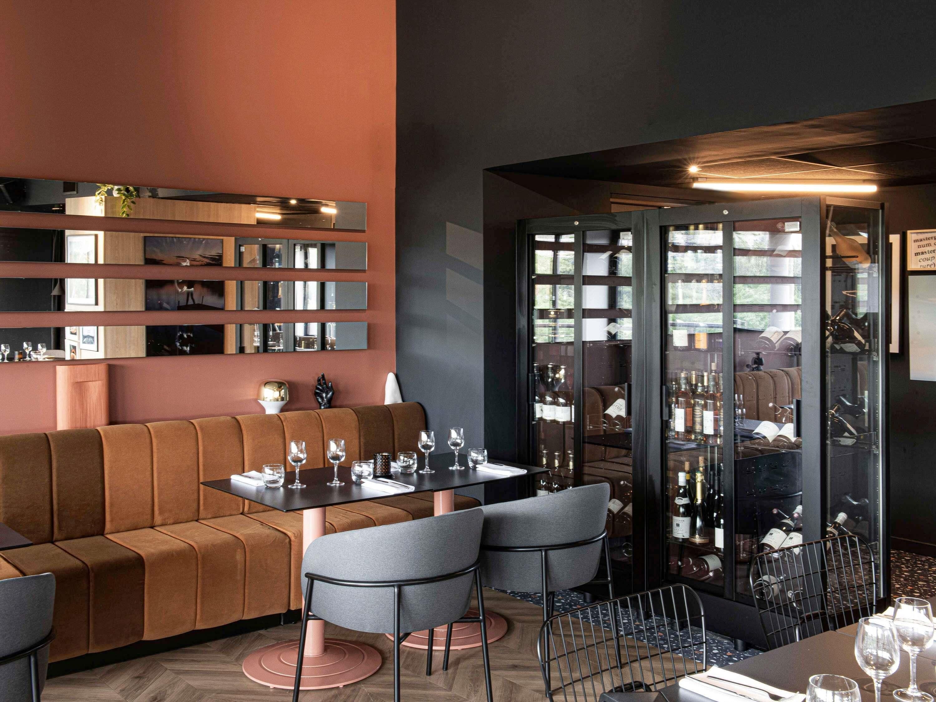 A modern restaurant interior with a brown sofa, grey chairs, a dining table set with wine glasses, and a wine rack visible through a glass partition.