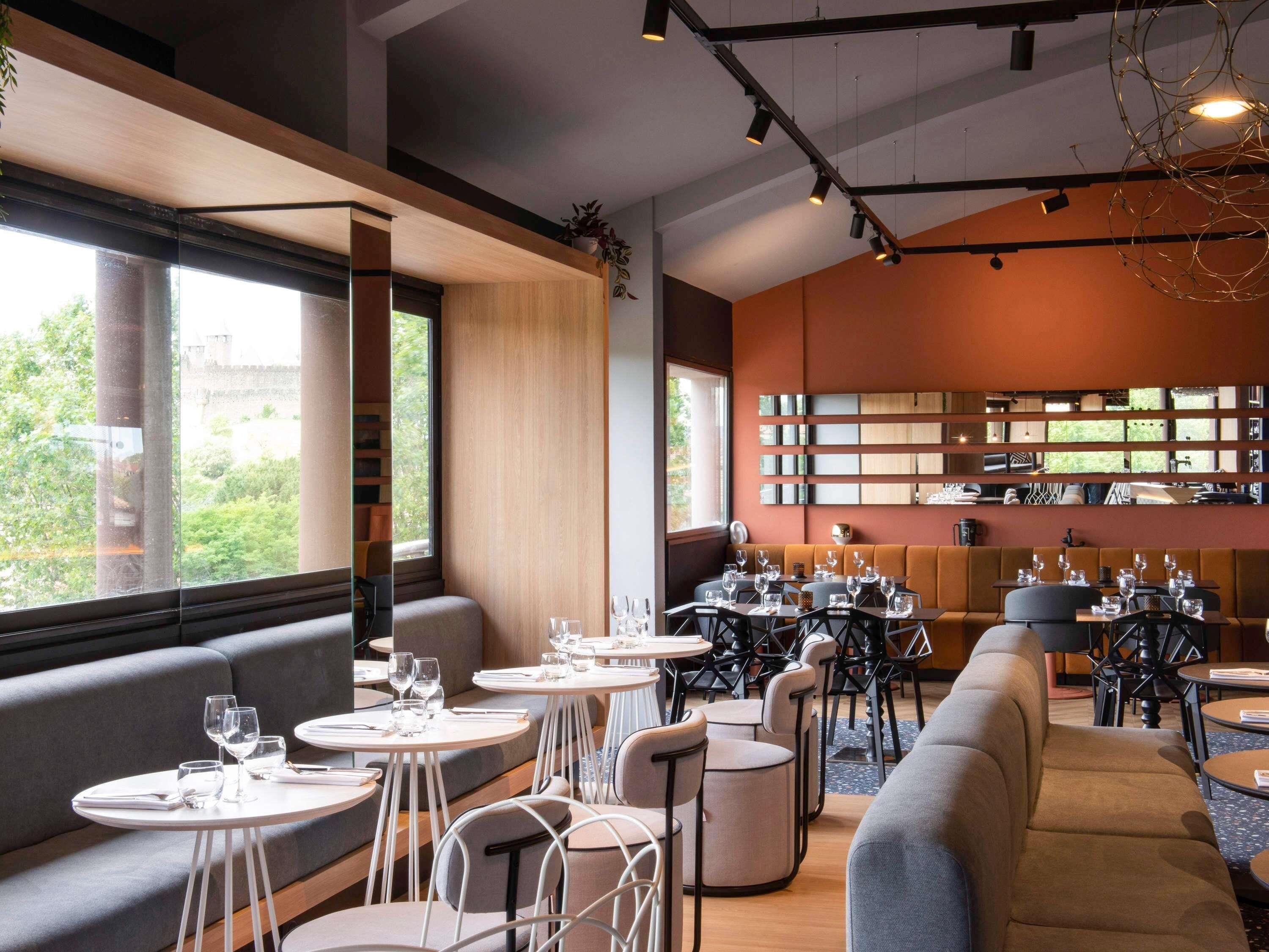 A modern restaurant with round tables, cushioned seating, and a view of greenery outside.