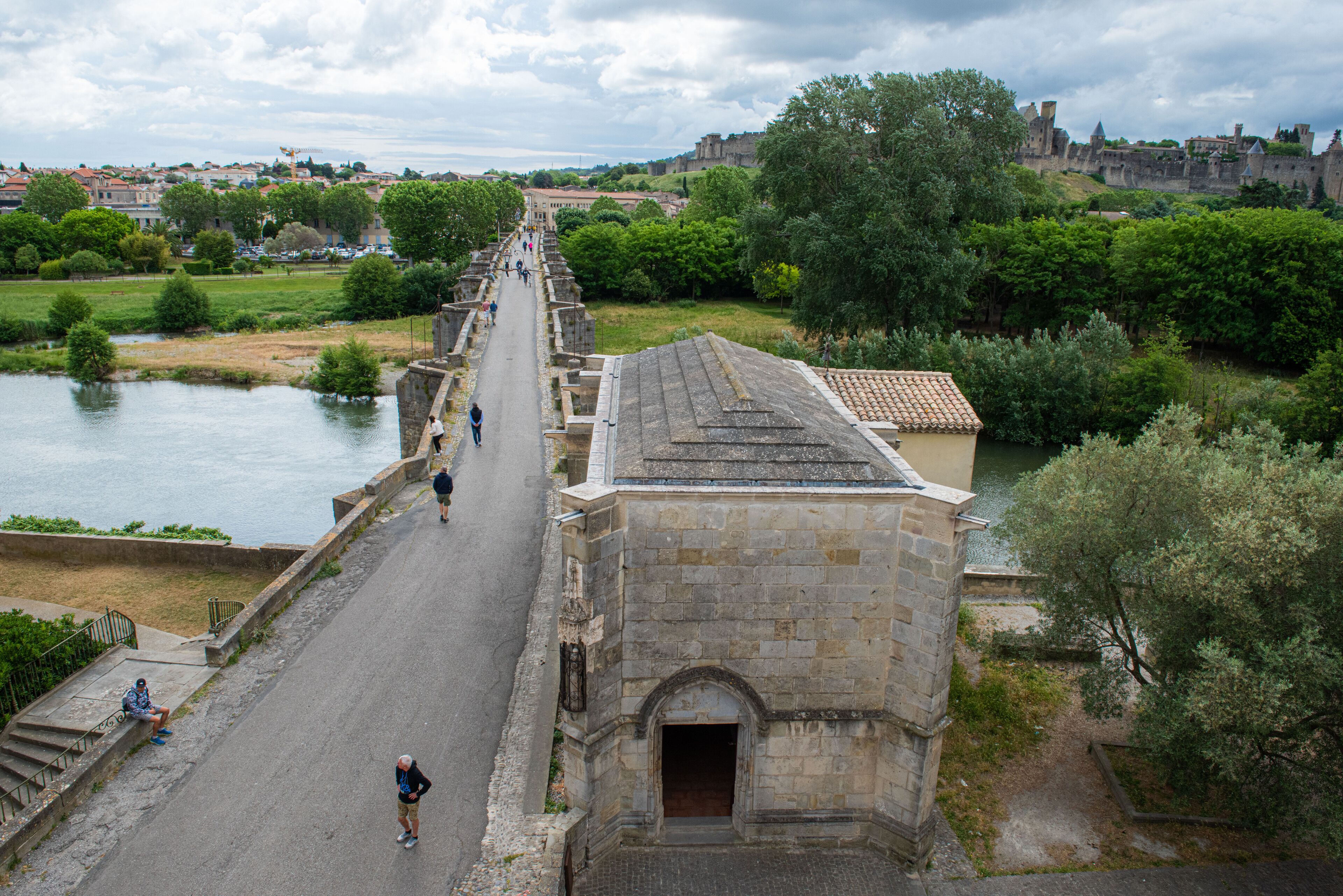 A stone bridge over a river with a pathway, surrounded by greenery and historic buildings.
