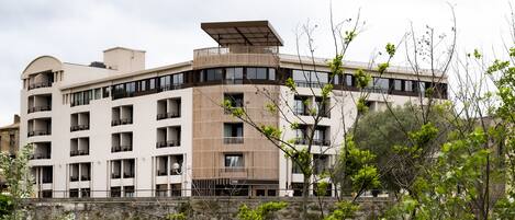 Un hotel moderno de varios pisos con balcones, un muro de piedra de contención y abundante vegetación en primer plano.
