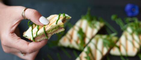 Una mano sostiene un sándwich con guarnición verde, junto a otros sándwiches y una flor sobre una superficie negra.