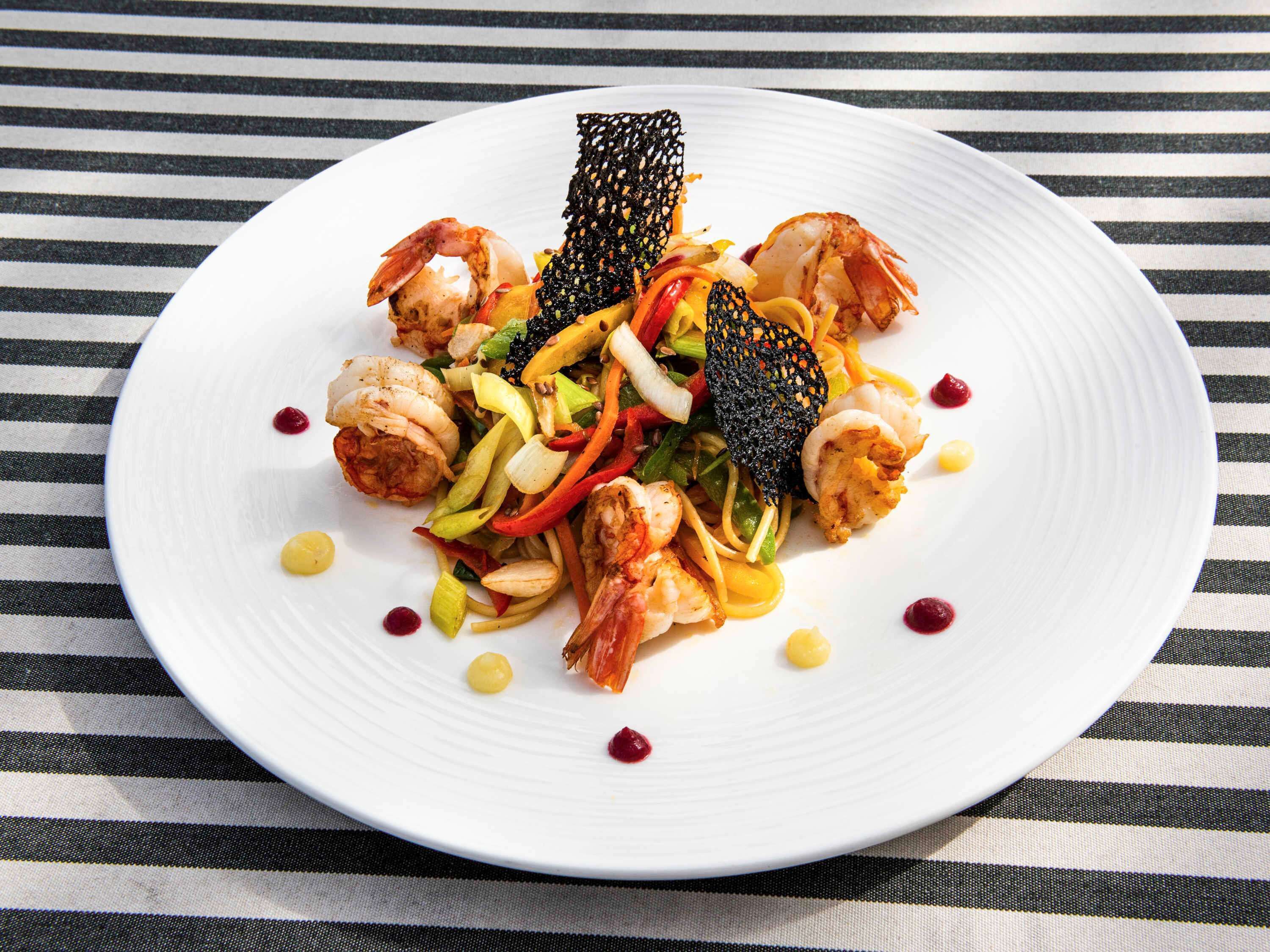 A white plate with a seafood dish, including shrimp and vegetables, on a striped fabric background.
