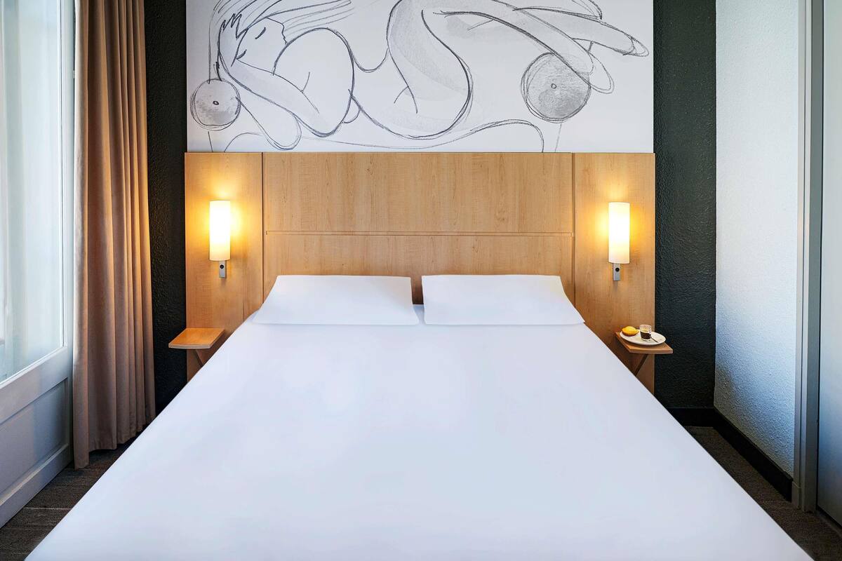 Habitación de hotel con una cama grande, dos mesitas de noche, una obra de arte en la pared y una ventana con cortinas.