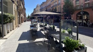 Un café al aire libre con mesas y sillas, rodeado de edificios con balcones y escaparates.