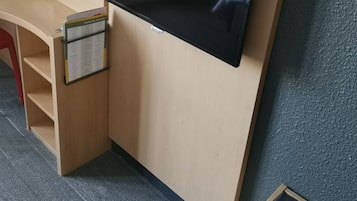 A hotel room with a flat-screen TV mounted on a wooden wall, a desk with a telephone, and a small seating area with a cushion.