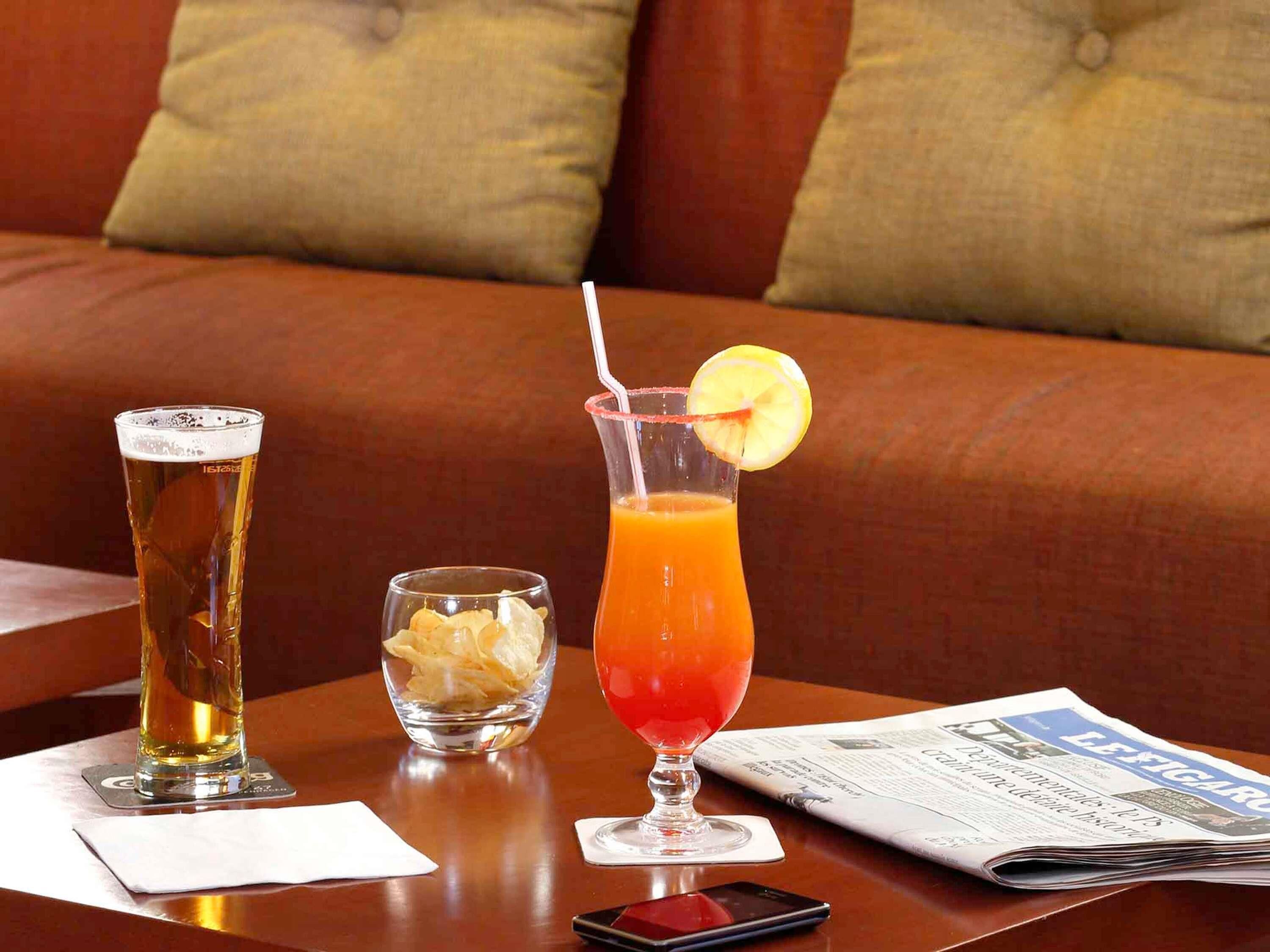 Ein Tisch mit zwei Getränken, einer kleinen Schale Chips, einer Zeitung und einem Smartphone.