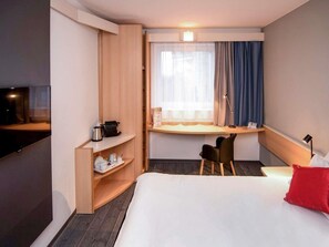 Een hotelkamer met een bed, een bureau met stoel, een televisie en een plank met spullen.