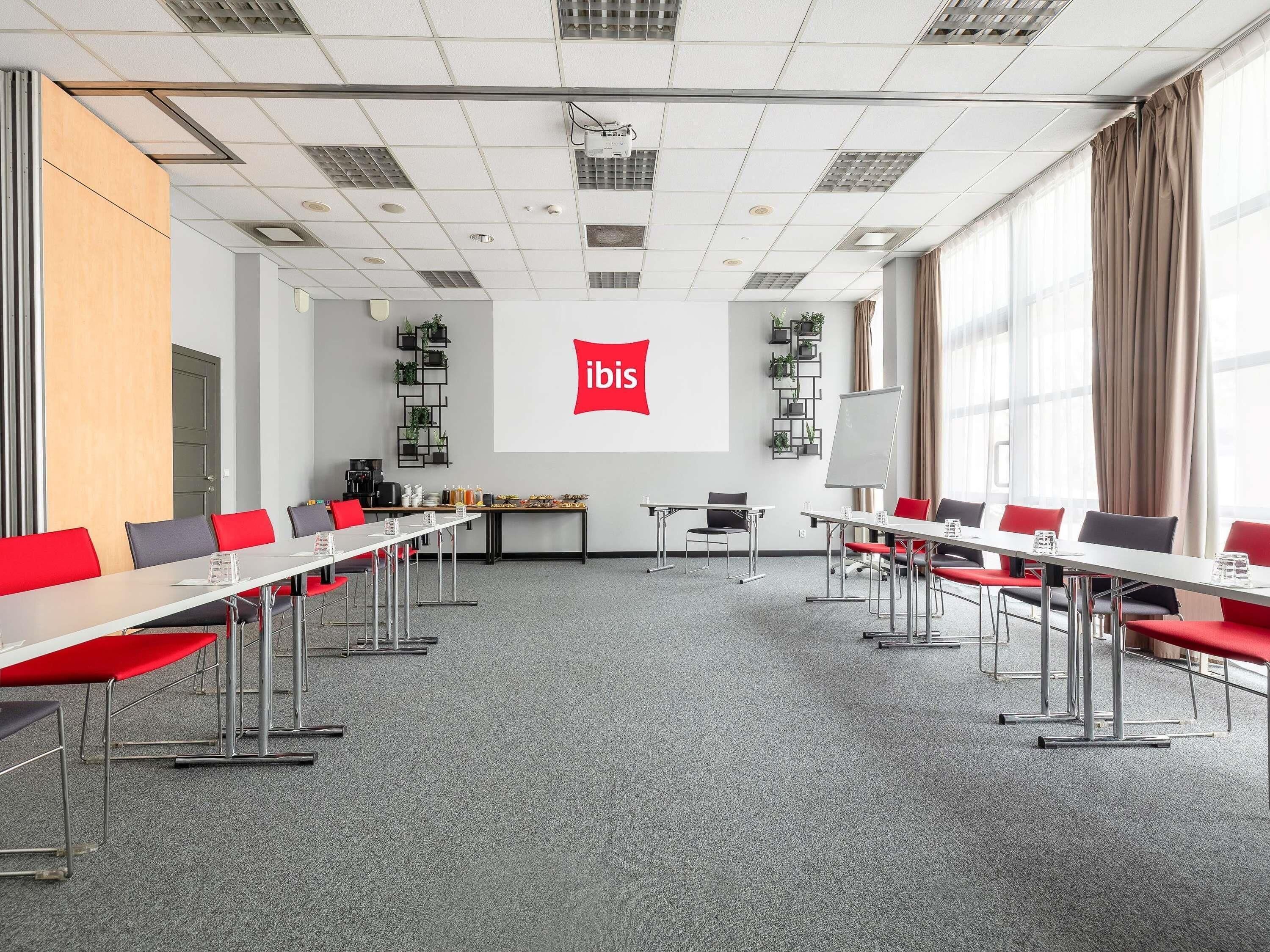A conference room with a projector screen displaying 'ibis,' tables with chairs, a coffee station, and a large window with curtains.