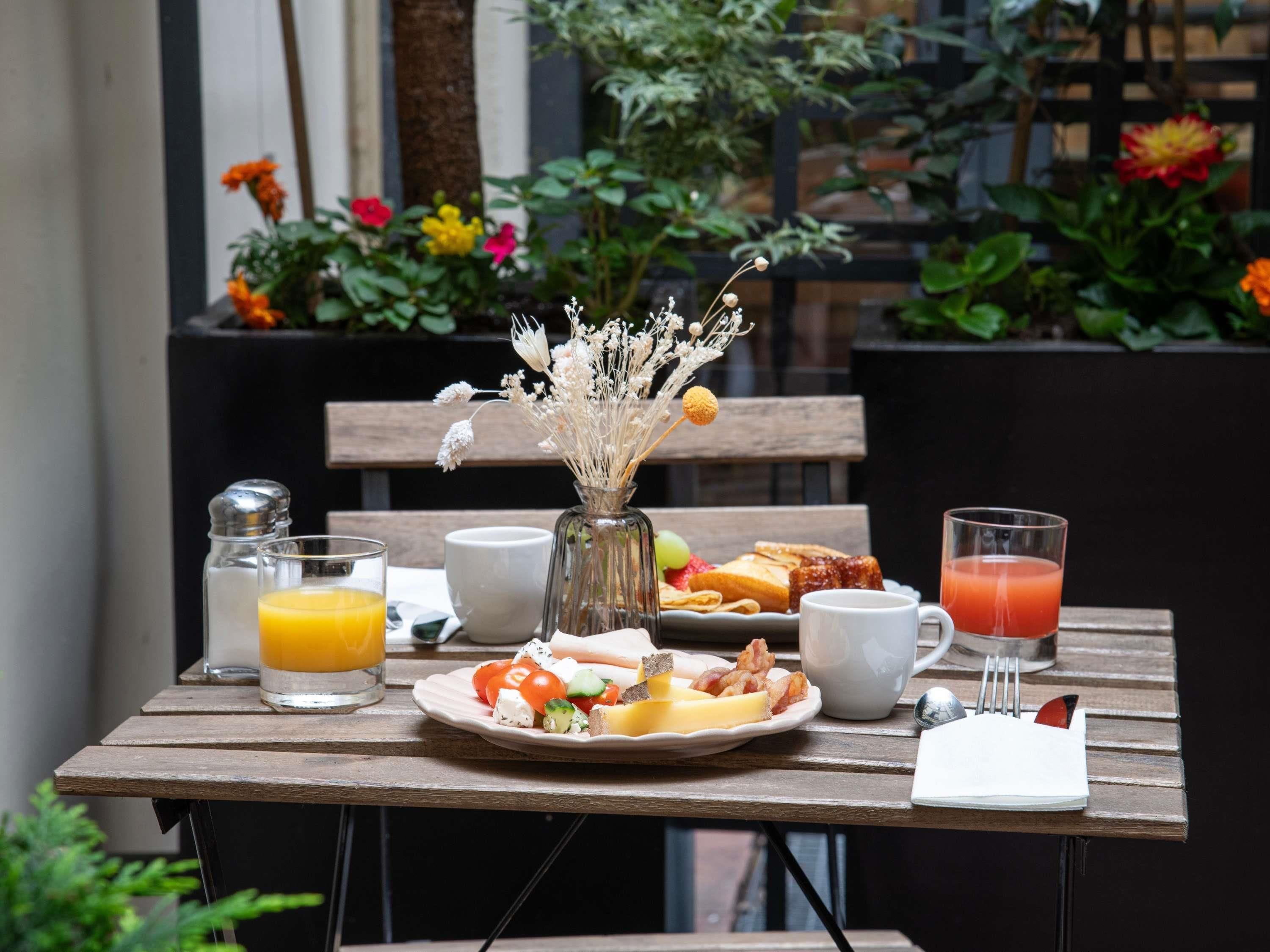 Ein Holztisch mit Frühstücksaufstellung, darunter eine Speisenplatte, ein Glas Orangensaft, ein Glas Tomatensaft, ein Salzstreuer, eine weiße Tasse und ein kleiner Blumenstrauß mit getrockneten Blumen.