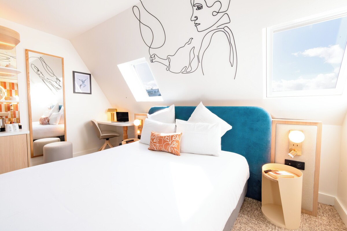 Moderni hotellihuone, jossa on sänky, työpöytä, tuoli, pieni pöytä ja yöpöytä.