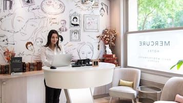 Eine Frau sitzt an einem Schreibtisch mit einem Laptop, umgeben von Büchern und Dekoration, in einem Raum mit einem großen Fenster und einer Wand mit künstlerischen Zeichnungen.