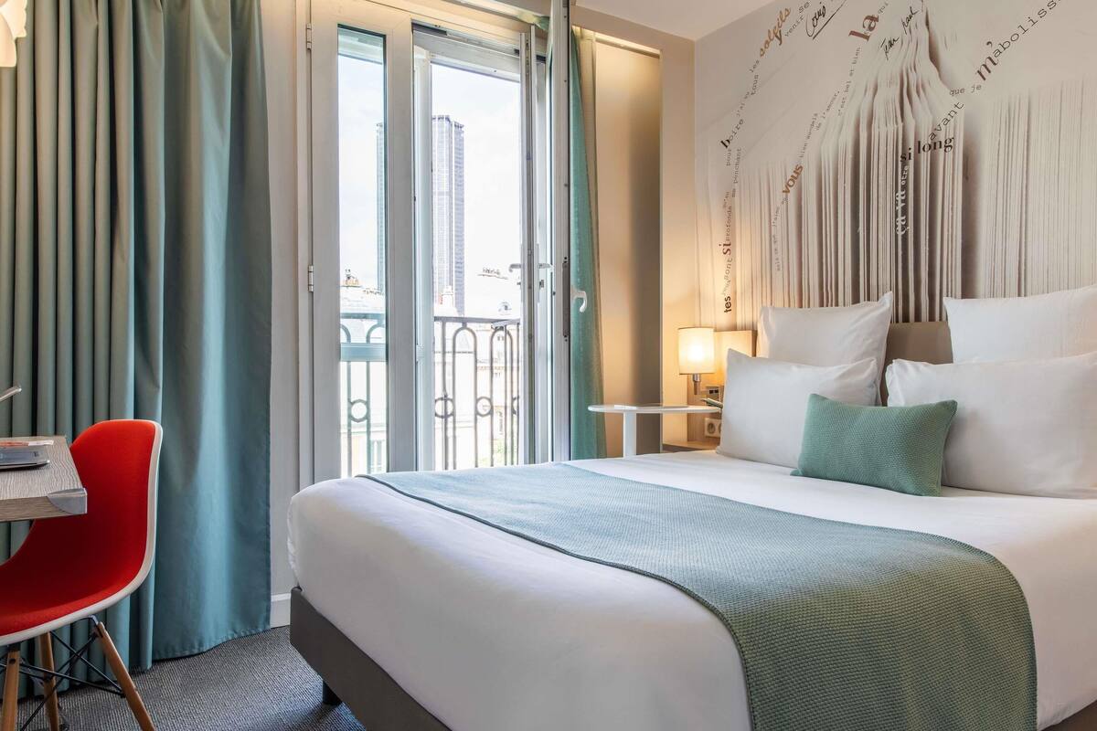 A hotel room with a bed, a desk, a chair, and a view of the city through the window.