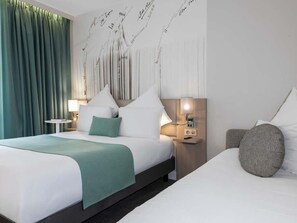 A hotel room with a bed, a bedside table, a lamp, and a wall with decorative elements. - Mercure Paris Montparnasse Raspail (Paris)