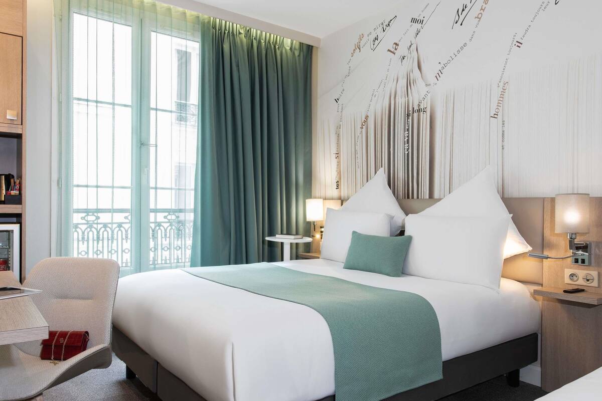 A hotel room with a bed, a chair, a table, and a window with curtains.