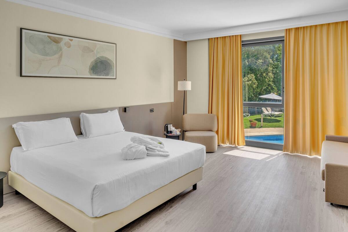 A hotel room with a large bed, a chair, a desk, and a view of a pool and greenery.
