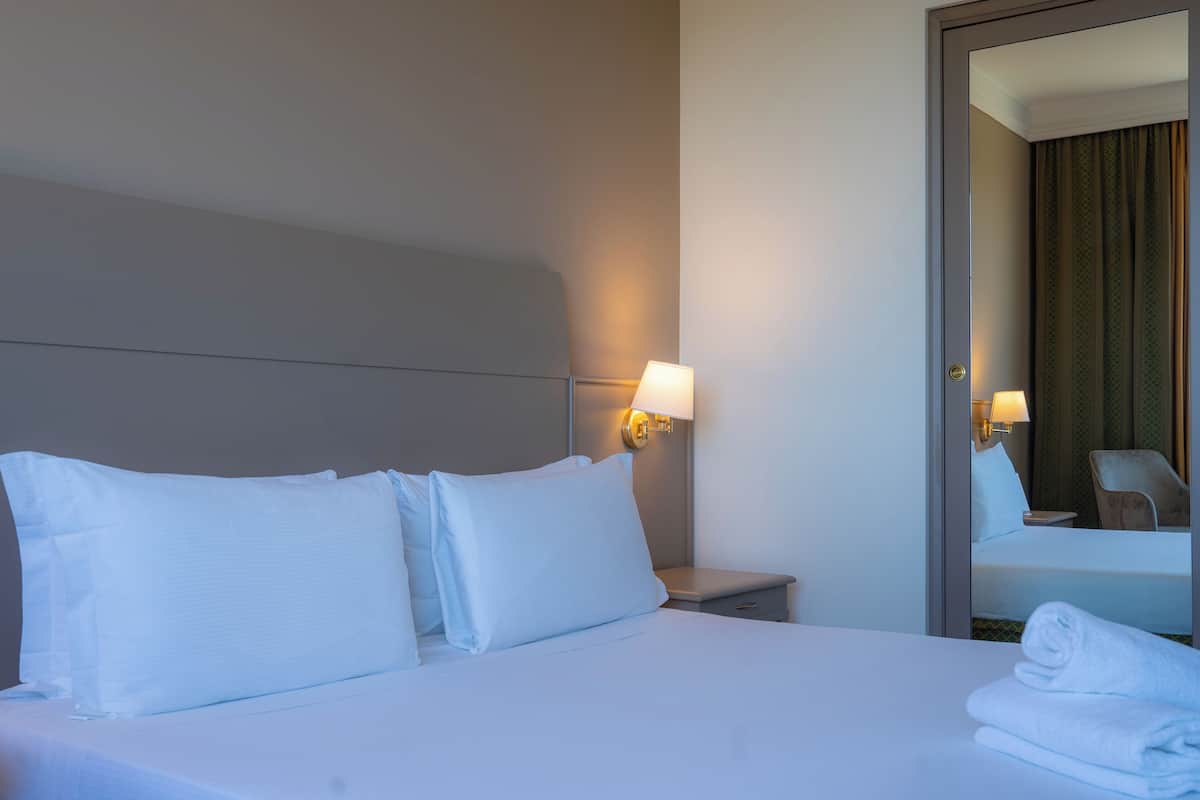 A hotel room with a neatly made bed, a bedside lamp, a nightstand, and a mirror reflecting the room.