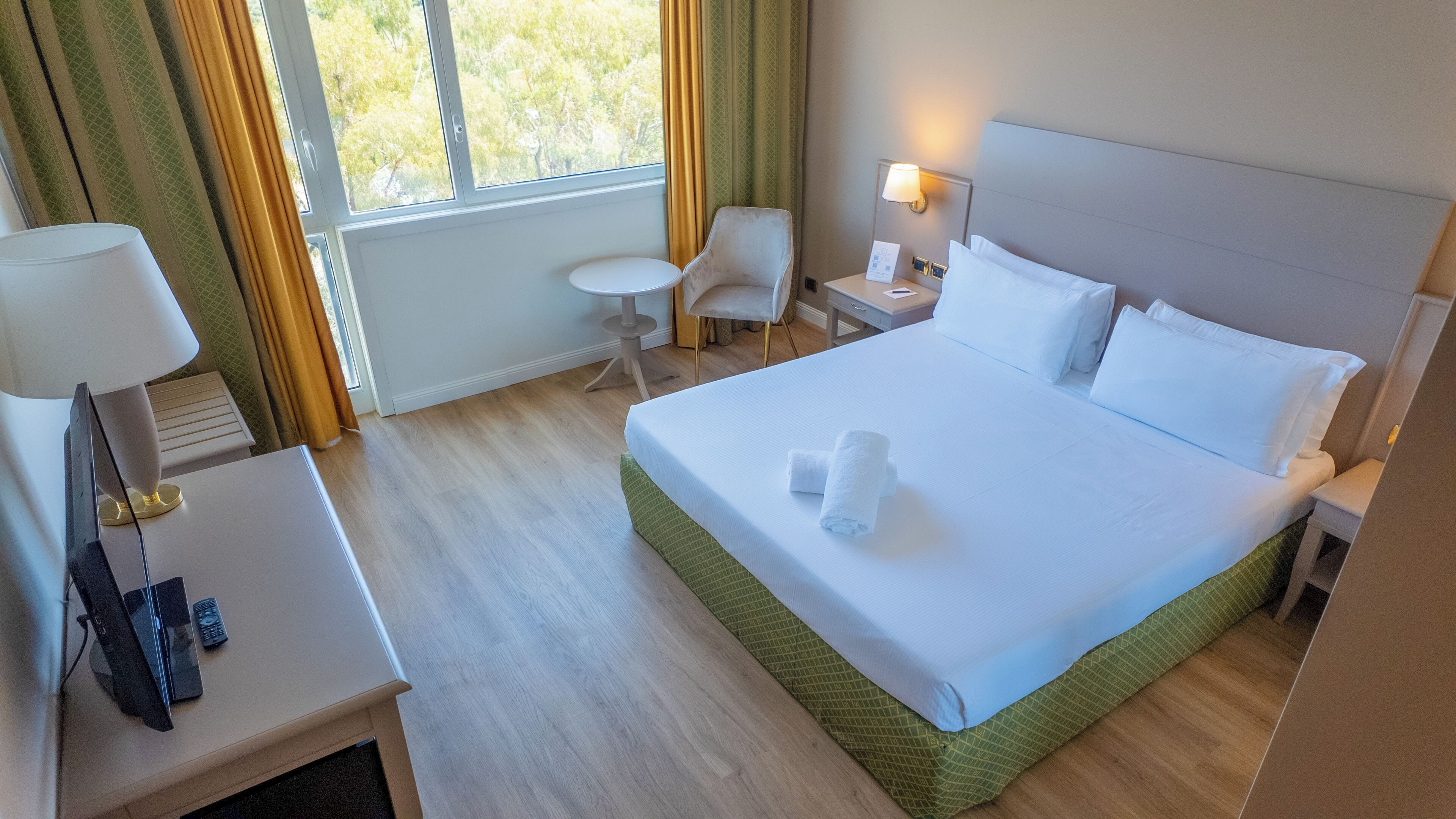 A hotel room with a large bed, a small table with a lamp, a chair, and a window with curtains.