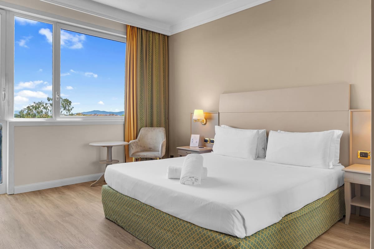A hotel room with a large bed, a chair, a small table, and a window with curtains.