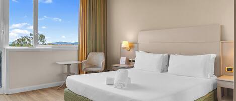 A hotel room with a large bed, a chair, a small table, and a window with curtains.