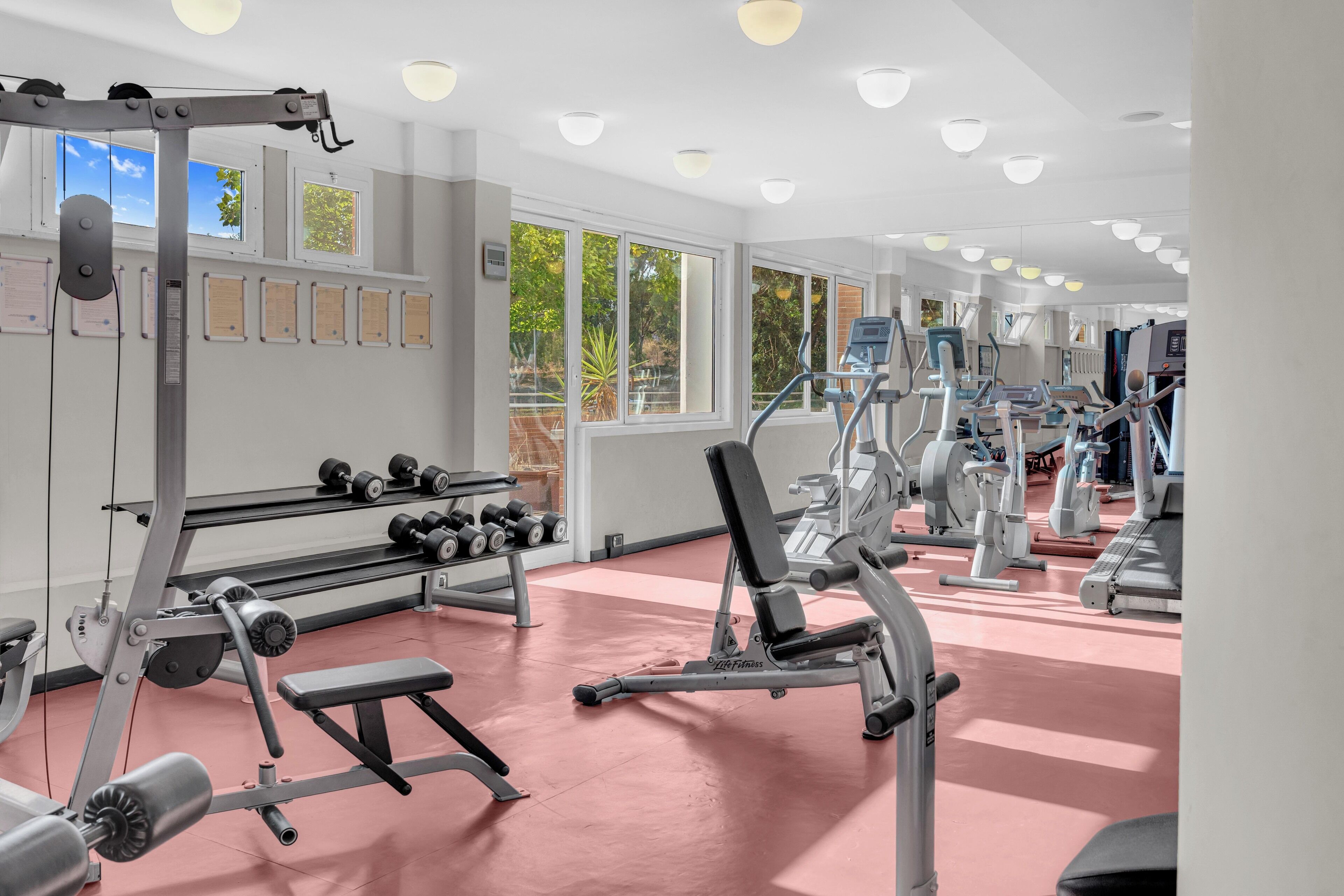Una palestra con diverse attrezzature per esercizi, ampie finestre e pavimento rosso.
