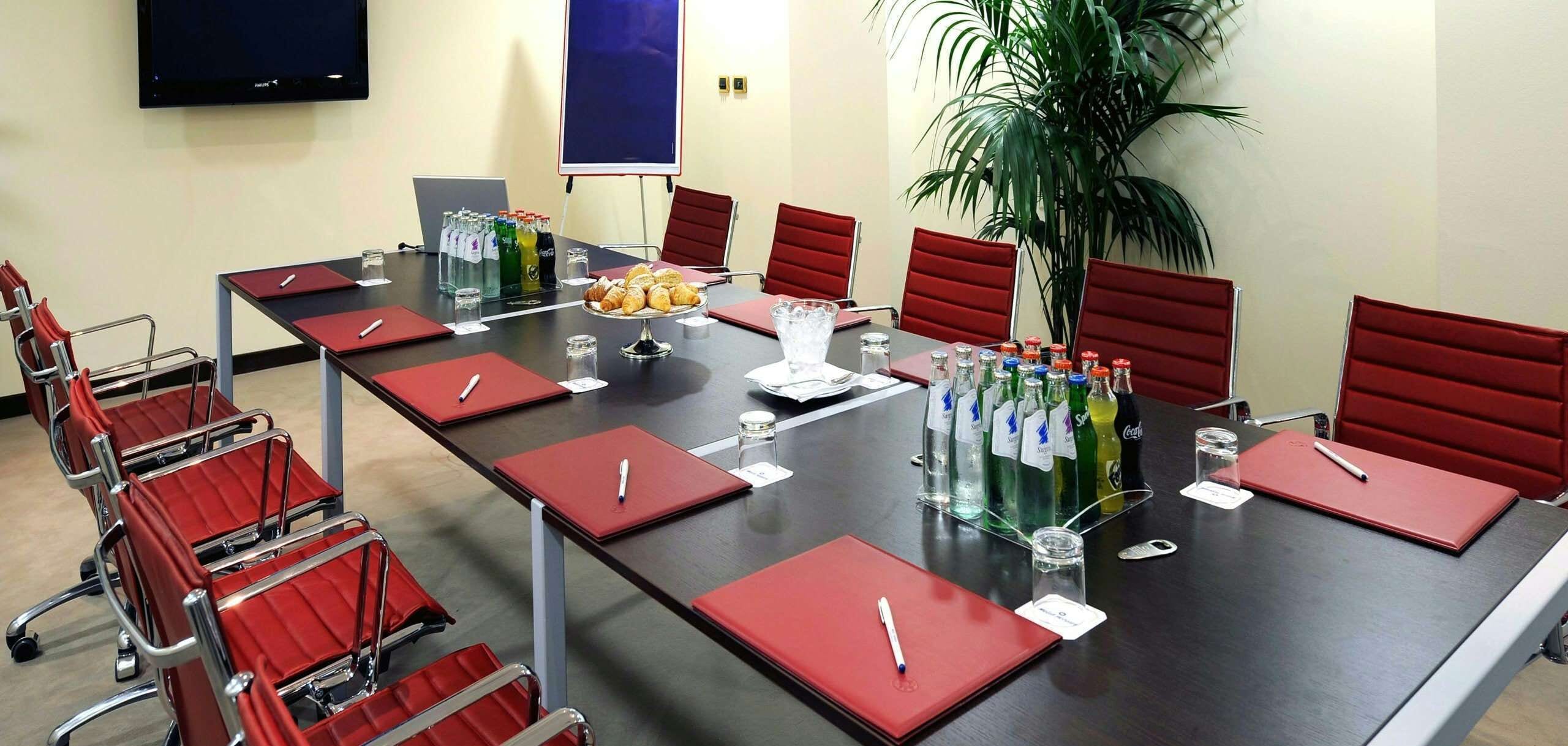 A conference room with a long table, red chairs, a laptop, a projector screen, and a potted plant.
